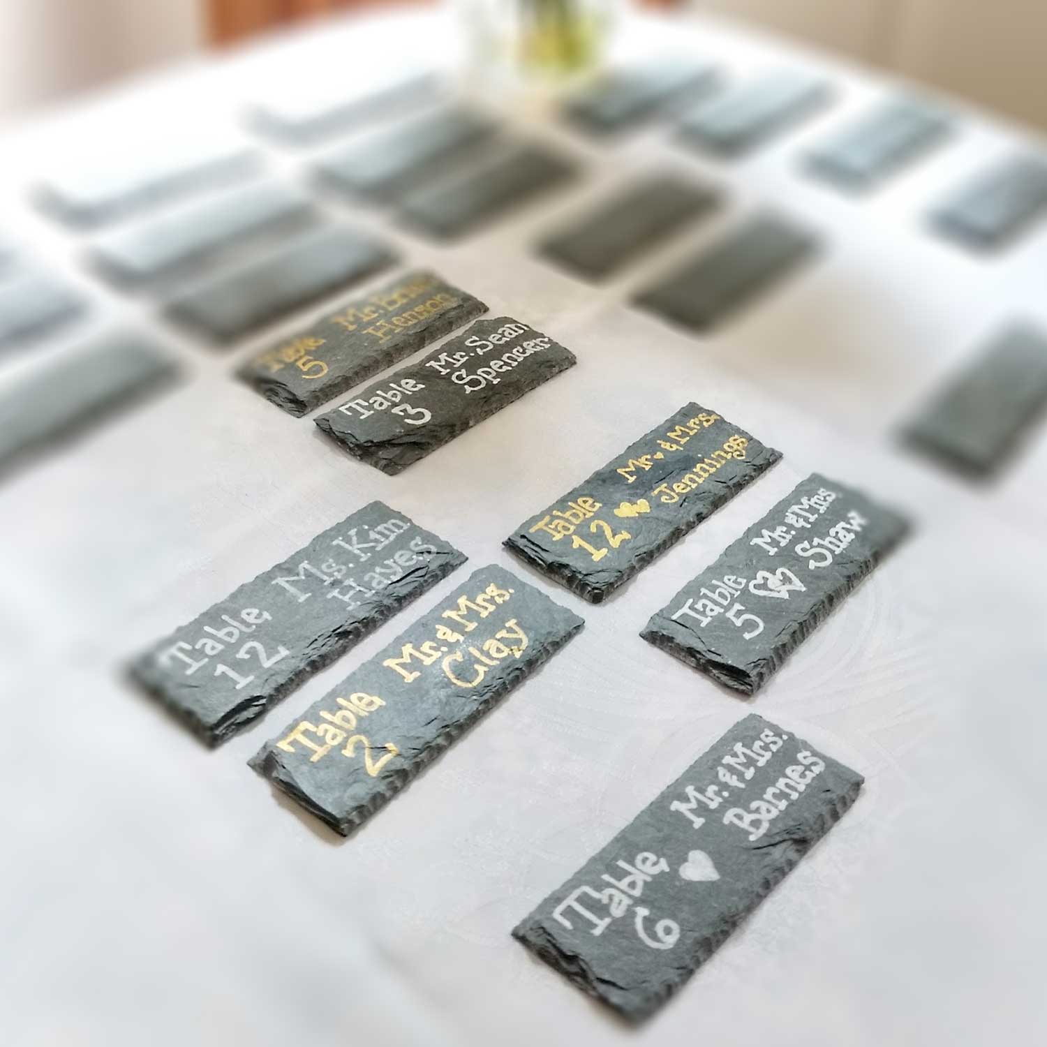 Slate wedding and event place cards on a white table cloth, each with names and table assignments written out.